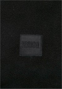 Un tissu noir à texture douce présente une étiquette rectangulaire noire embossée avec "Urban Classics" dans un design minimaliste.