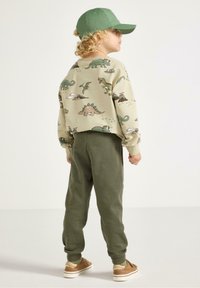 Beiger Pullover mit Dinosaurier-Print, grüne Jogginghosen und eine passende grüne Kappe. Weicher Stoff mit gerippten Bündchen und entspannter Passform. Braune Schuhe.