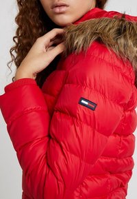 Kvinde i en lys rød jakke med dun, med hætte beklædt med pels, der justerer kraven nær hagen, hvilket viser et lille rektangulært logo på ærmet.