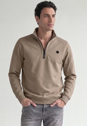 Uomo con capelli scuri e ricci che indossa un maglione beige con zip parziale e jeans grigi, in piedi con le mani in tasca, guardando a destra.