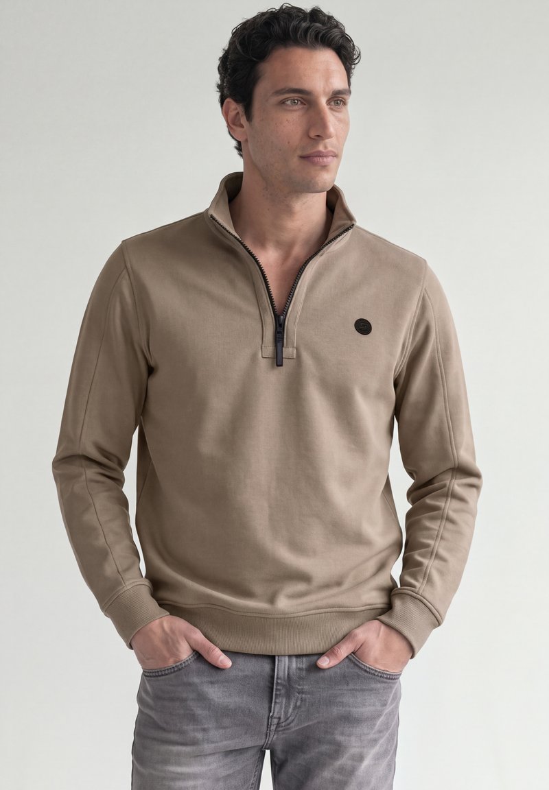 Homme aux cheveux bouclés et foncés portant un pull beige à fermeture éclair quart et un jean gris, debout les mains dans les poches, regardant vers la droite.