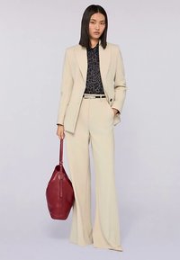 Beige op maat gemaakt pak met wijde pijpen, zwarte blouse met patroon en een dieprode leren handtas. Kenmerkt zich door een gestructureerd ontwerp en strakke lijnen.