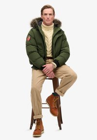 Grøn puffjakke med pelsfor på hætten, cremefarvet kabelstrikket sweater, beige bukser og brune snørebåndsstøvler, siddende på en træskammel.