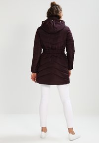 Lila vadderad jacka med huva, åtskuren midja och chevron-sticksöm. Buren över vita byxor och vita skor, sedd bakifrån.