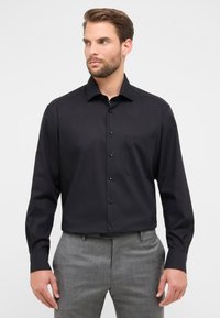 Camicia nera a maniche lunghe con colletto a punta, caratterizzata da una tasca sul petto e una consistenza liscia. Abbinata a pantaloni grigi.