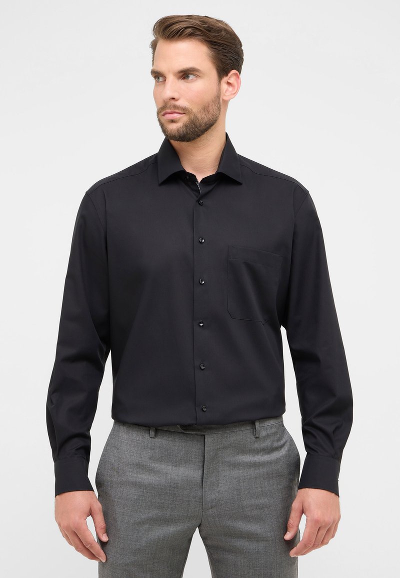 Camicia nera a maniche lunghe con colletto a punta, caratterizzata da una tasca sul petto e una consistenza liscia. Abbinata a pantaloni grigi.