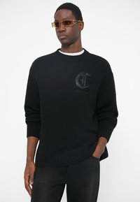 Pulover negru tricotat cu guler rotund, având mansete și tiv cu benzi. Include un detaliu subtil cu logo în relief pe piept.