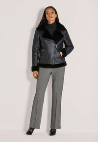 Zwarte imitatieleren jas met een bontkraag en manchetten, voorzien van een diagonale ritssluiting en twee voorkantzakken. Gecombineerd met patroonbroeken met wijde pijpen.