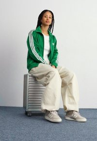 Grön sportjacka med vita ränder, vit t-shirt, beige lösa byxor och ljusgrå sneakers, sittande på en metallförvaringsenhet.