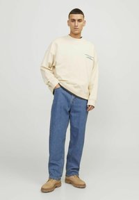 Mlad moški stoji s rokami v žepih, oblečen v bež pulover, modre kavbojke in svetlo rjave čipkaste čevlje, proti enostavni svetli ozadju.