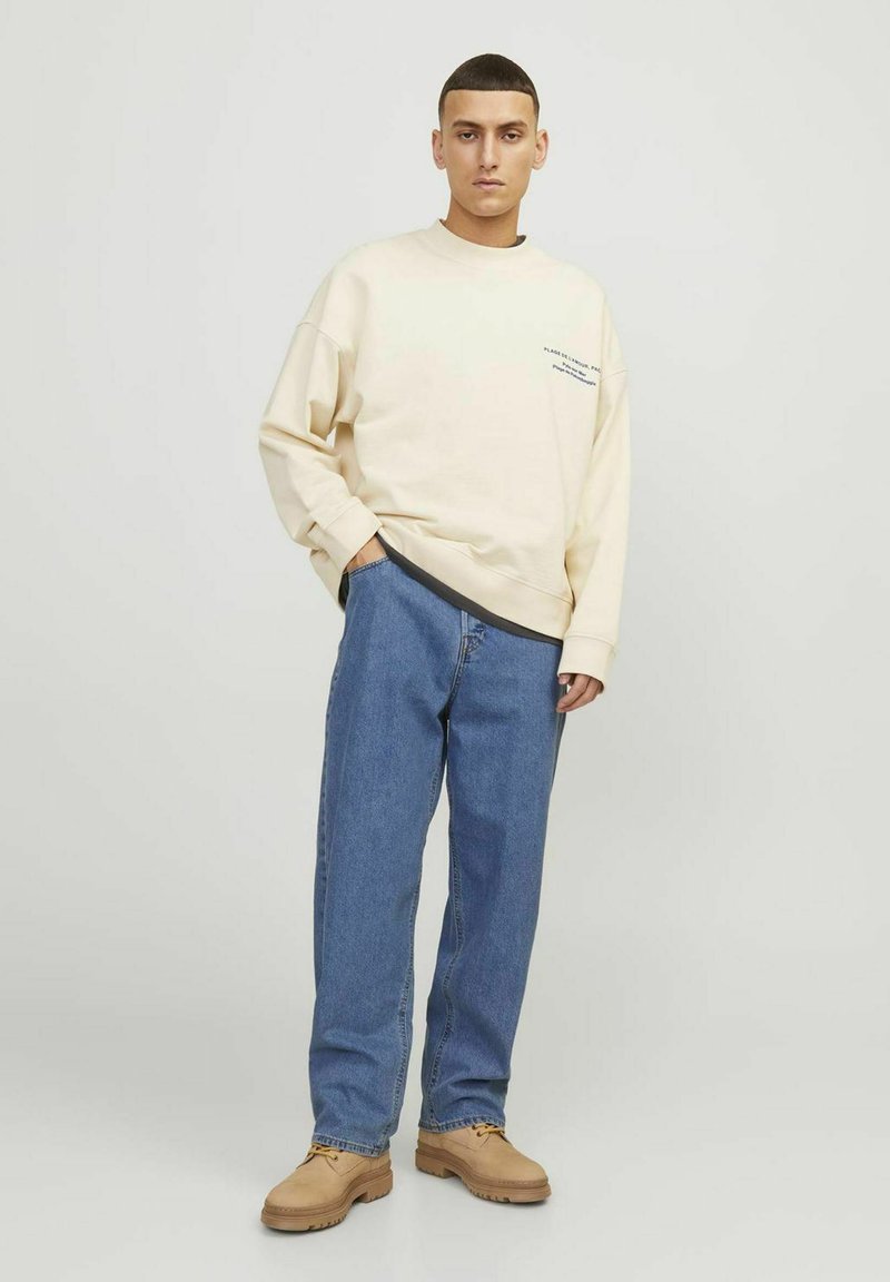 Młody mężczyzna stoi z rękami w kieszeniach, ubrany w beżowy sweter, niebieskie dżinsy i brązowe buty z sznurówkami na tle jednolitego jasnego tła.