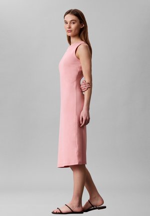 Kvinde stående i profil i en ærmeløs pink midi-kjole og sorte flade sandaler mod en ensfarvet grå baggrund.