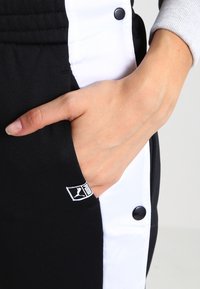Zwarte sportbroek met witte zijpanelen, voorzien van drukknoopjes en een gestructureerde stof. Zichtbaar logo-label op de zak. Hand in de zak.