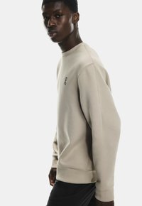 Hanorac bej cu guler rotund, manșete și tiv cu nervuri. Prezintă un mic logo negru pe piept. Material moale cu o textură netedă.