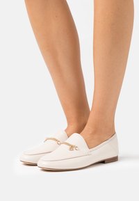 Mocassins en cuir blanc avec un talon bas, ornés d'une chaînette dorée sur le devant. Texture lisse, bout arrondi et design minimaliste.