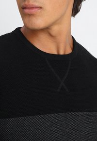 Maglione nero lavorato a maglia con design strutturato, caratterizzato da una scollatura rotonda e una fascia orizzontale grigia a contrasto lungo il bordo inferiore.
