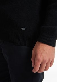 Maglione nero a maglia con polsini e orlo a coste, caratterizzato da una piccola etichetta logata argento sul lato. Il materiale sembra morbido e testurizzato.