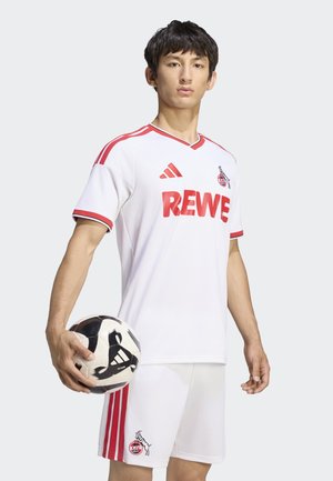 Ung mand iført hvidt og rødt fodboldtøj holder en sort-hvid Adidas fodbold mod en ensfarvet baggrund.