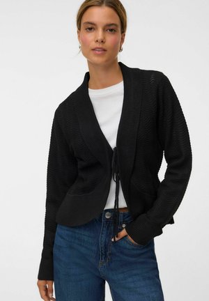 Kvinde iført en sort strikket cardigan over en hvid skjorte og blå jeans, stående med den ene hånd i lommen mod en ensfarvet baggrund.