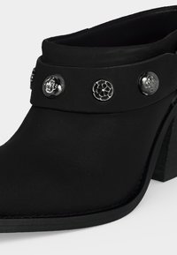 Mule noire à talon avec un bout arrondi, ornée de trois clous décoratifs métalliques sur la bride supérieure.