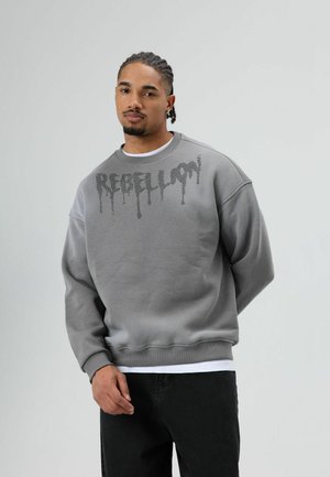 Grijze sweatshirts met doorgezakte schouders en geribbelde boorden, met het woord "REBELLION" in een druipend ontwerp over de borst.