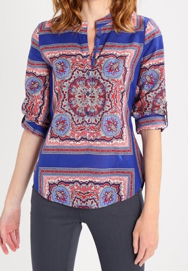 Femme portant un chemisier bleu à motifs avec les manches retroussées et un pantalon foncé sur un fond uni.
