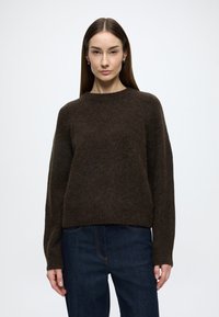 Barna gyapjúkeverék pulóver hosszú ujjakkal, kerek nyakkivágással és laza szabással, sötét farmernadrággal párosítva. Puha, texturált felület részletekkel.