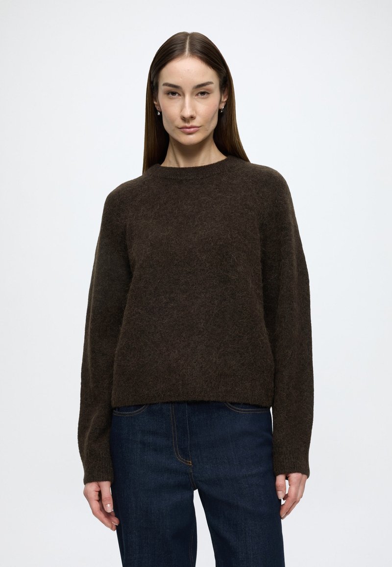 Barna gyapjúkeverék pulóver hosszú ujjakkal, kerek nyakkivágással és laza szabással, sötét farmernadrággal párosítva. Puha, texturált felület részletekkel.