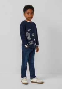 Marineblauwe sweatshirt met witte graphics, lange mouwen en gestreepte manchetten, gecombineerd met blauwe jeans en witte sneakers met tan details.