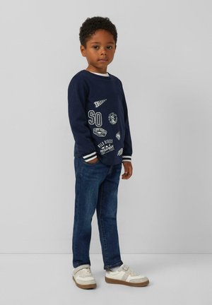 Marineblauwe sweatshirt met witte graphics, lange mouwen en gestreepte manchetten, gecombineerd met blauwe jeans en witte sneakers met tan details.
