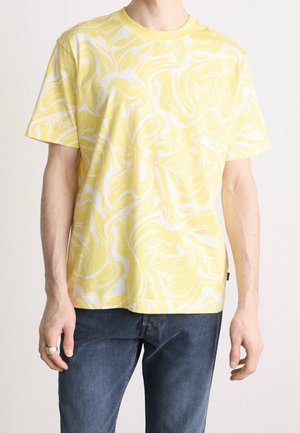 Hombre vestido con una camiseta de manga corta a rayas amarillas y blancas y jeans de color azul oscuro, de pie con los brazos relajados a los lados.