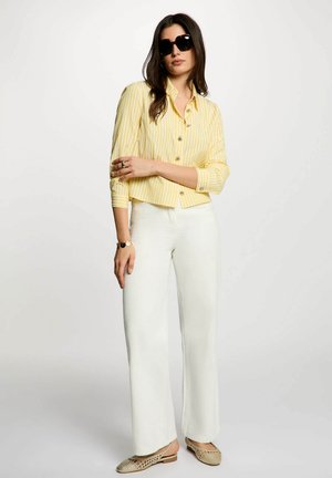 Donna con grandi occhiali da sole neri, camicia gialla a righe con bottoni, pantaloni bianchi a gamba larga e ballerine beige intrecciate, in piedi con le braccia incrociate.