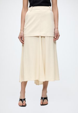 Žena nosí vrstvenú krémovú midi sukňu so štruktúrovanou hornou vrstvou a čiernymi sandálmi na podpätku.