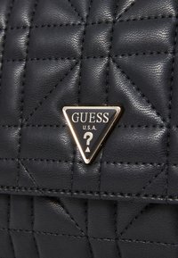 Schwarze gesteppte Handtasche mit glänzender Oberfläche, featuring ein dreieckiges GUESS-Logo in Gold und Schwarz. Die Nähte bilden ein Musterdesign.