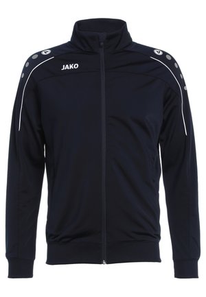 Veste de sport zippée marine en tissu lisse, avec des accents blancs et des boutons ronds sur les épaules. Logo "JAKO" sur la poitrine.