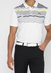 Polo blanc avec une partie supérieure rayée grise et jaune à motifs, arborant un petit logo. Porté avec un pantalon noir et une ceinture blanche.