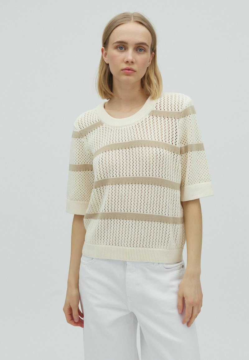 Jeune femme aux cheveux blonds porte un pull en maille à manches courtes rayé beige et blanc et un pantalon blanc sur un fond uni.