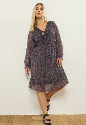 Femme portant une robe midi à manches longues à motif avec un ourlet à volants, des sandales noires et un collier pendentif en or, debout contre un mur uni.