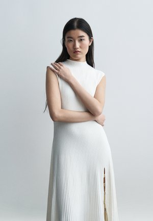 Jeune femme aux longs cheveux foncés portant une robe blanche côtelée sans manches avec une fente latérale, debout les bras croisés devant un fond uni.