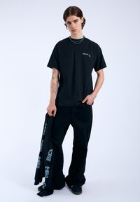 Camiseta de algodón negra con un pequeño logo blanco en el pecho, combinada con pantalones de pierna ancha negros que presentan impresiones gráficas en azul claro a lo largo de una pierna.
