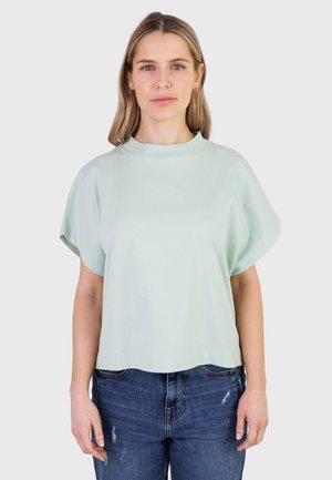 Kvinde med skulderlangt lyst blondt hår iført en lys mintfarvet oversized kortærmet top og blå slidte jeans, stående mod en ensfarvet baggrund.