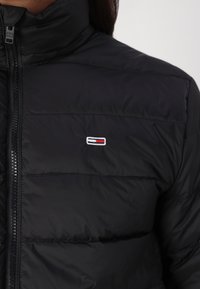 Zwarte pufferjack met een hoge kraag, horizontale stiksels en een klein geborduurd logo op de borst. Zichtbare ritsluiting.