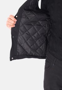 Giacca nera con zip, con un interno in satin trapuntato, caratterizzata da chiusure a scatto e polsini a costine. Il materiale esterno presenta un motivo testurizzato.