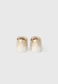 Beige sneakers med en tekstureret overdel, der har et glat guldaccent på hælen og en hvid gummisål. Rund form, lav-top design.