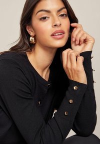 Svart stickad tröja med rund halsringning, med detaljer i guldknappar på ärmarna. Slät textur och figurnära design.