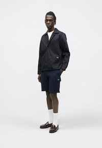 Chaqueta ligera azul marino con bolsillo, combinada con pantalones cortos azul marino que cuentan con un bolsillo parche. La modelo lleva calcetines blancos y mocasines oscuros.