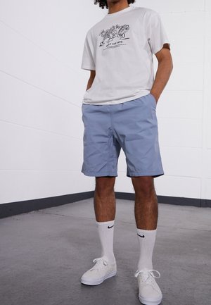 Mann trägt ein weißes Grafik-T-Shirt, hellblaue Shorts, weiße Nike Socken und weiße Sneakers und steht mit den Händen in den Taschen.