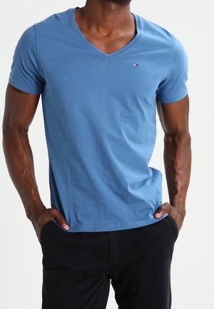 Homme portant un t-shirt bleu ajusté à col en V et manches courtes, avec un pantalon noir, les mains dans les poches, sur un fond blanc.