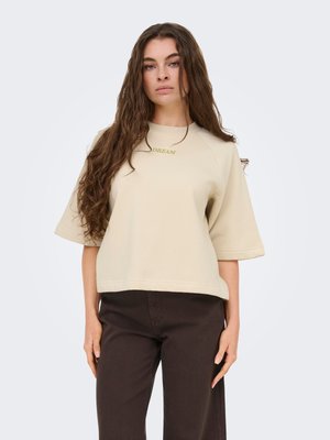 Femme aux longs cheveux bruns portant un haut beige à manches courtes avec le texte « DREAM » et un pantalon marron foncé, debout devant un fond clair uni.