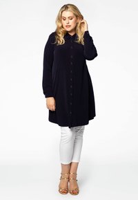 Marineblauwe knop-up tuniek met lange mouwen, voorzien van een klassieke kraag en een losse pasvorm. Gecombineerd met witte cropped pantalon en gouden sandaaltjes met banden.